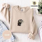 Sand crewneck sweatshirt folded with denim shorts, white sneakers and a potted plant, embroidered with a river otter head in brown and cream thread with long golden whiskers