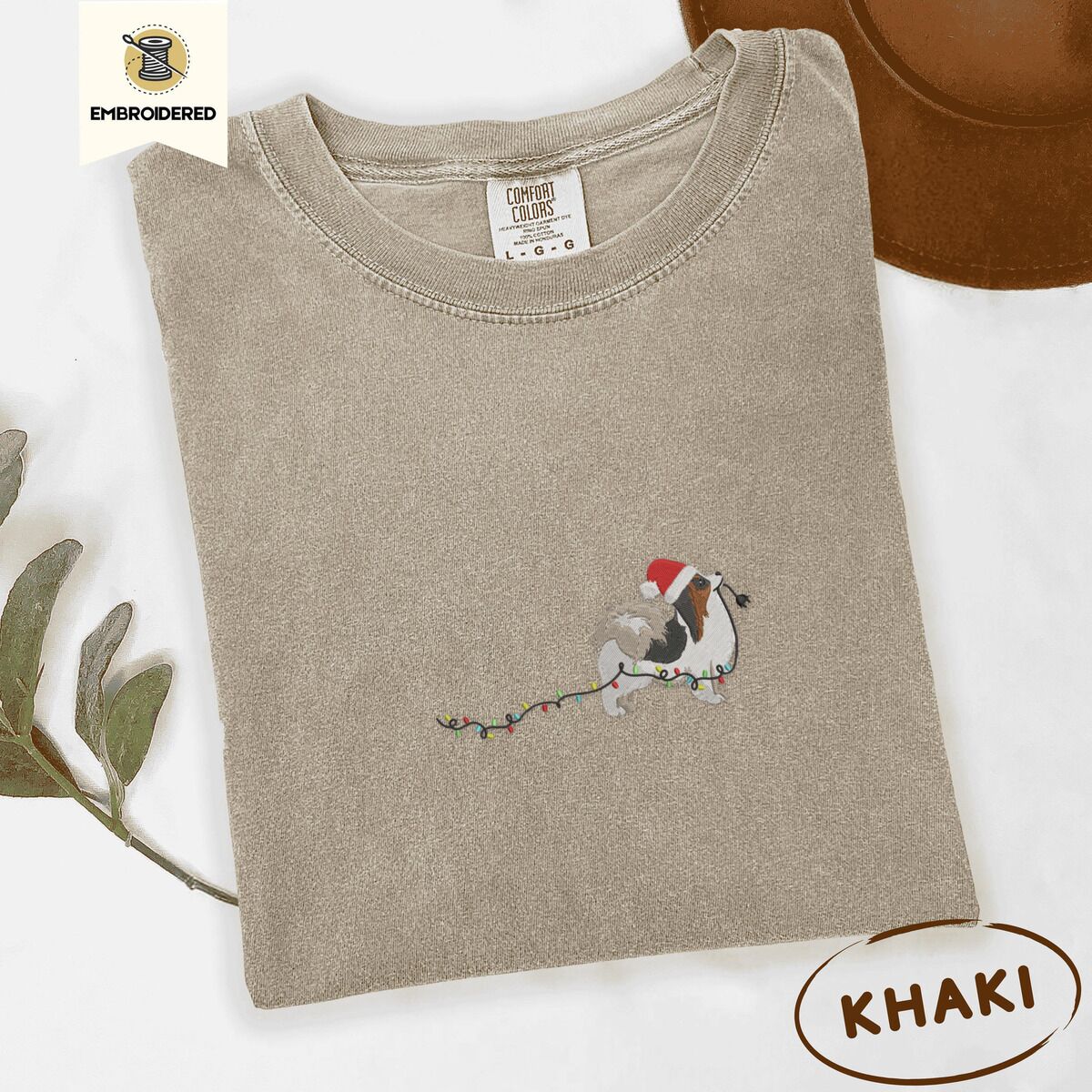 Papillon Christmas Lights Shirt – Khaki Khaki Comfort Colors T-shirt embroidered with a tricolor Papillon in a Santa hat holding the plug of a string of Christmas lights in its mouth