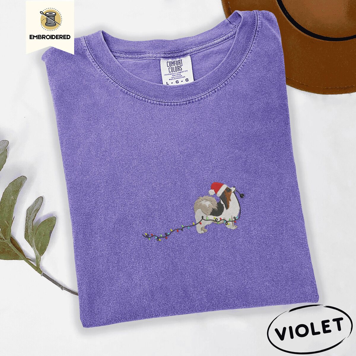 Papillon Christmas Lights Shirt – Violet Violet Comfort Colors T-shirt embroidered with a tricolor Papillon in a Santa hat holding the plug of a string of Christmas lights in its mouth