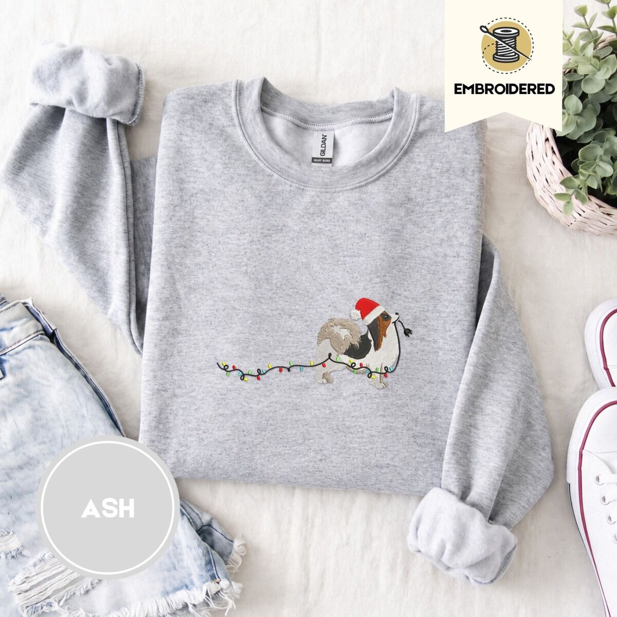 Papillon Christmas Sweatshirt – Ash Ash crewneck sweatshirt folded with denim shorts, white sneakers and a potted plant, embroidered with a tricolor Papillon in a Santa hat holding the plug of a string of Christmas lights in its mouth