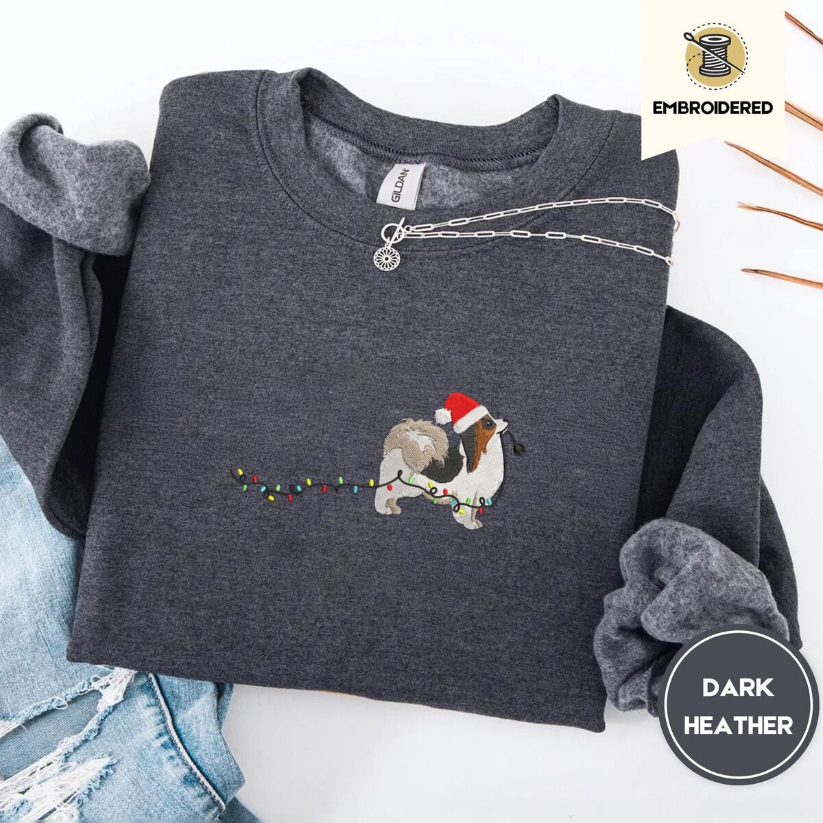 Papillon Christmas Sweatshirt – Dark Heather Dark Heather crewneck sweatshirt folded with ripped jeans and a silver chain necklace, embroidered with a tricolor Papillon in a Santa hat holding the plug of a string of Christmas lights in its mouth
