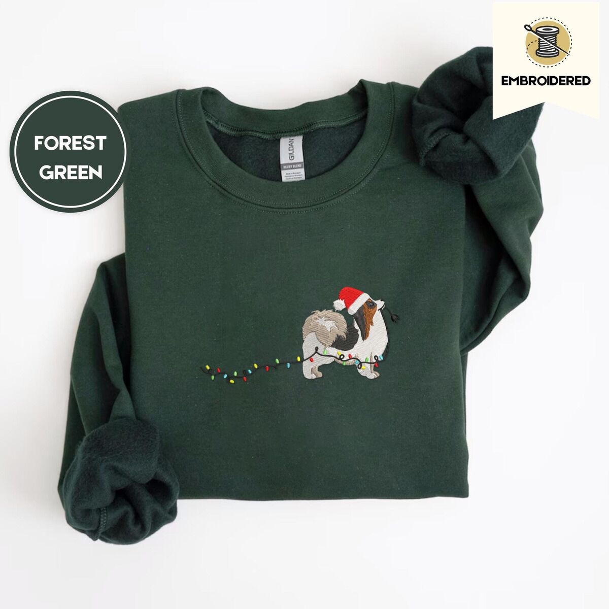 Papillon Christmas Sweatshirt – Forest Green Forest Green crewneck sweatshirt folded on a white background, embroidered with a tricolor Papillon in a Santa hat holding the plug of a string of Christmas lights in its mouth