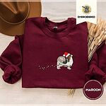 Maroon crewneck sweatshirt with a brown felt hat and dried bunny tail grass, embroidered with a tricolor Papillon in a Santa hat holding the plug of a string of Christmas lights in its mouth