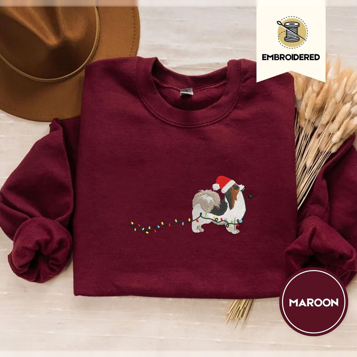 Papillon Christmas Sweatshirt – Maroon Maroon crewneck sweatshirt with a brown felt hat and dried bunny tail grass, embroidered with a tricolor Papillon in a Santa hat holding the plug of a string of Christmas lights in its mouth