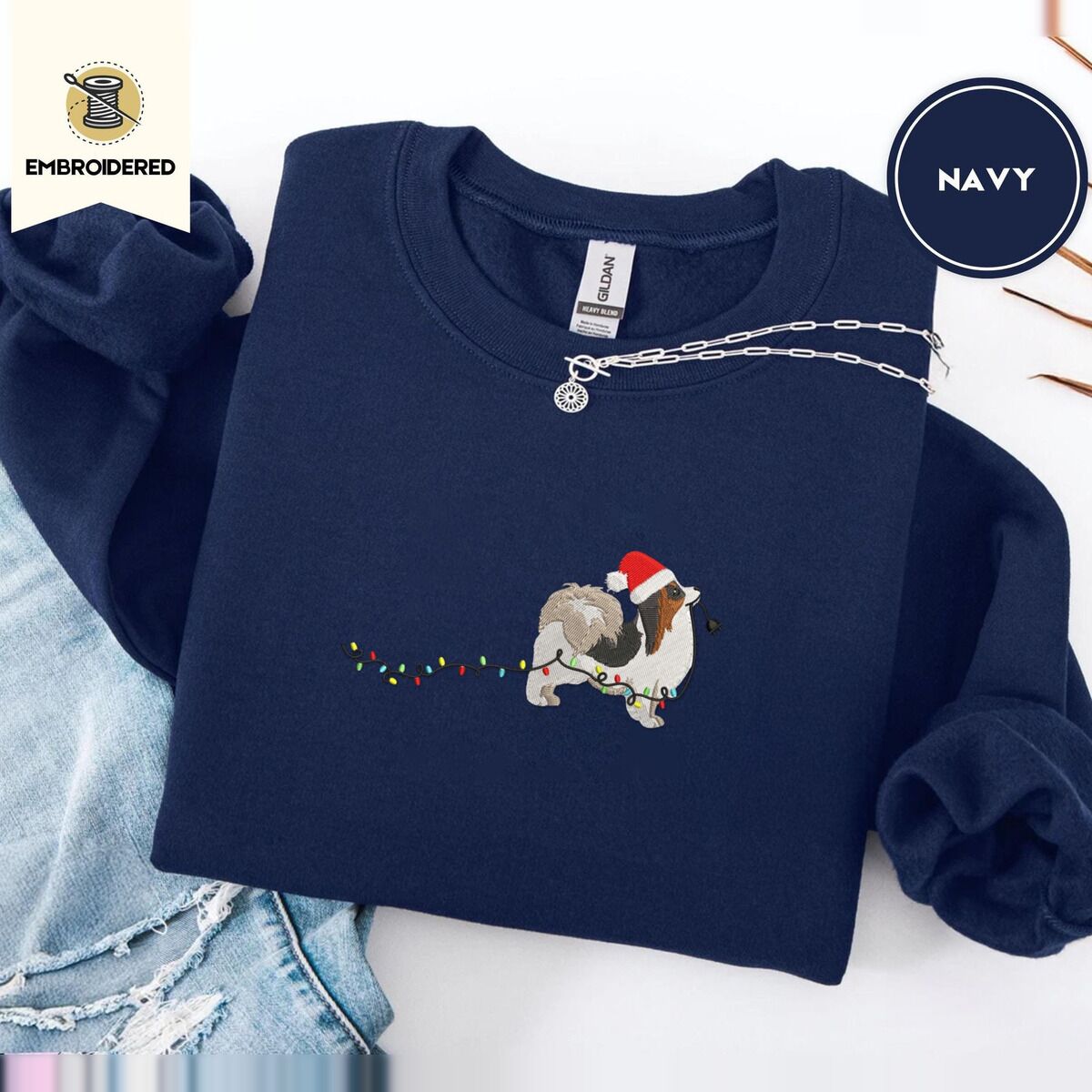 Papillon Christmas Sweatshirt – Navy Navy crewneck sweatshirt folded with ripped jeans and a silver chain necklace, embroidered with a tricolor Papillon in a Santa hat holding the plug of a string of Christmas lights in its mouth