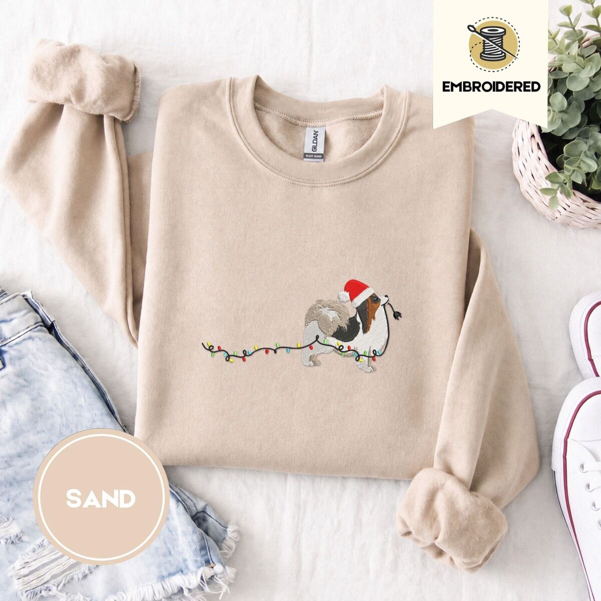 Papillon Christmas Sweatshirt – Sand Sand crewneck sweatshirt folded with denim shorts, white sneakers and a potted plant, embroidered with a tricolor Papillon in a Santa hat holding the plug of a string of Christmas lights in its mouth