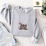 Ash crewneck sweatshirt folded with denim shorts, white sneakers and a potted plant, embroidered with a fluffy brown tabby kitten with big round eyes peeking over a ledge at a pink butterfly