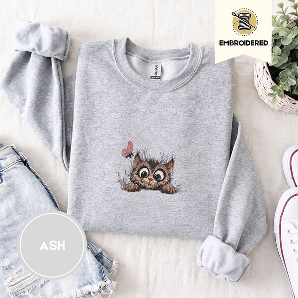 Ash crewneck sweatshirt folded with denim shorts, white sneakers and a potted plant, embroidered with a fluffy brown tabby kitten with big round eyes peeking over a ledge at a pink butterfly