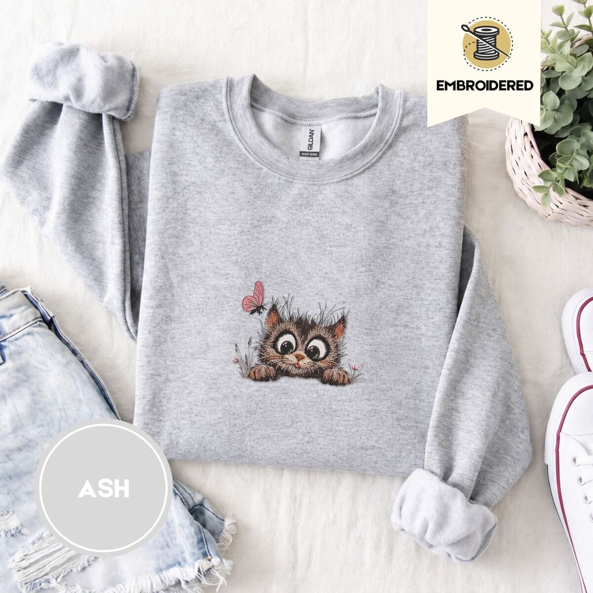 Peeking Cat Sweatshirt – Ash Ash crewneck sweatshirt folded with denim shorts, white sneakers and a potted plant, embroidered with a fluffy brown tabby kitten with big round eyes peeking over a ledge at a pink butterfly