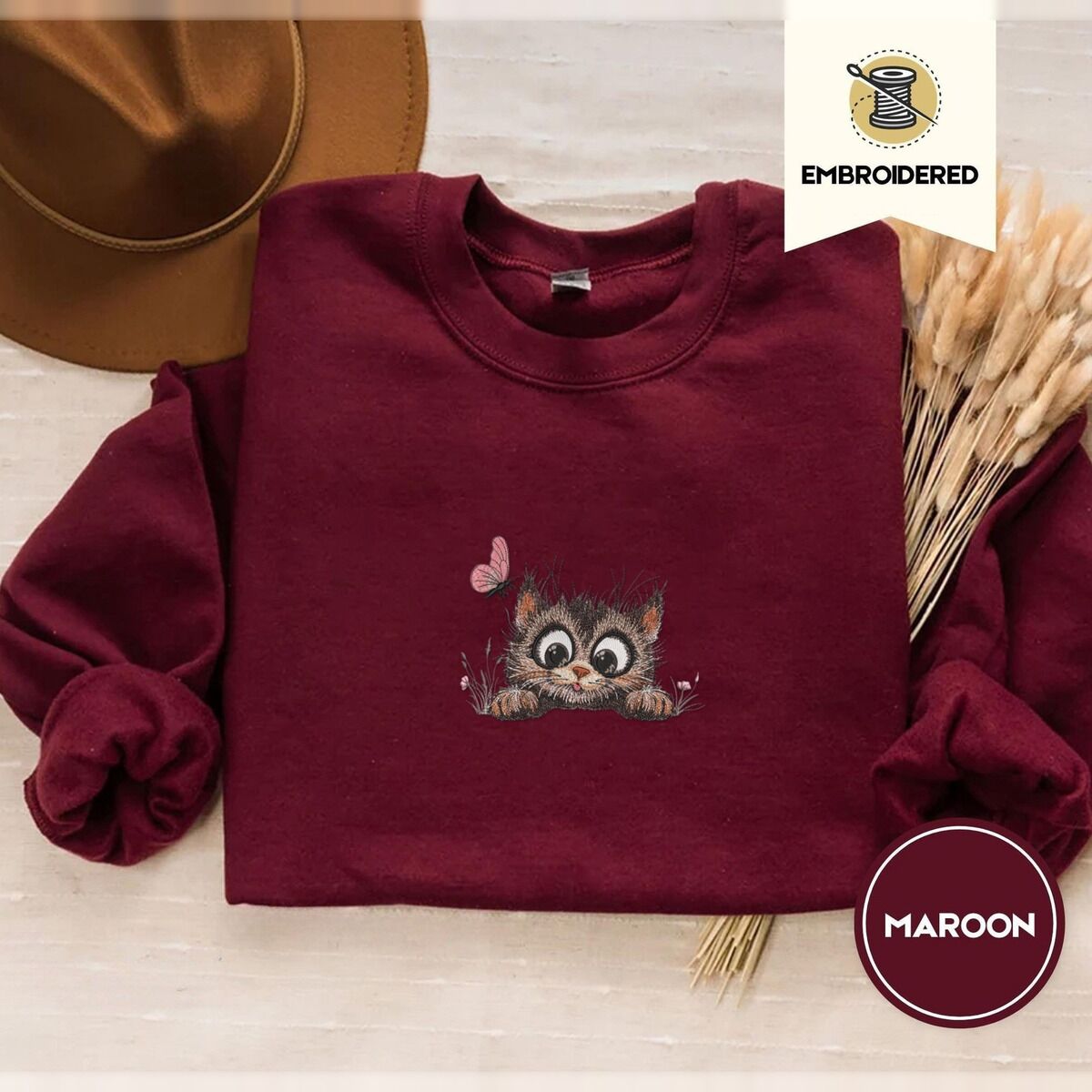 Peeking Cat Sweatshirt – Maroon Maroon crewneck sweatshirt with a brown felt hat and dried bunny tail grass, embroidered with a fluffy brown tabby kitten with big round eyes peeking over a ledge at a pink butterfly