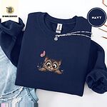 Navy crewneck sweatshirt folded with ripped jeans and a silver chain necklace, embroidered with a fluffy brown tabby kitten with big round eyes peeking over a ledge at a pink butterfly