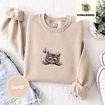 Sand crewneck sweatshirt folded with denim shorts, white sneakers and a potted plant, embroidered with a fluffy brown tabby kitten with big round eyes peeking over a ledge at a pink butterfly