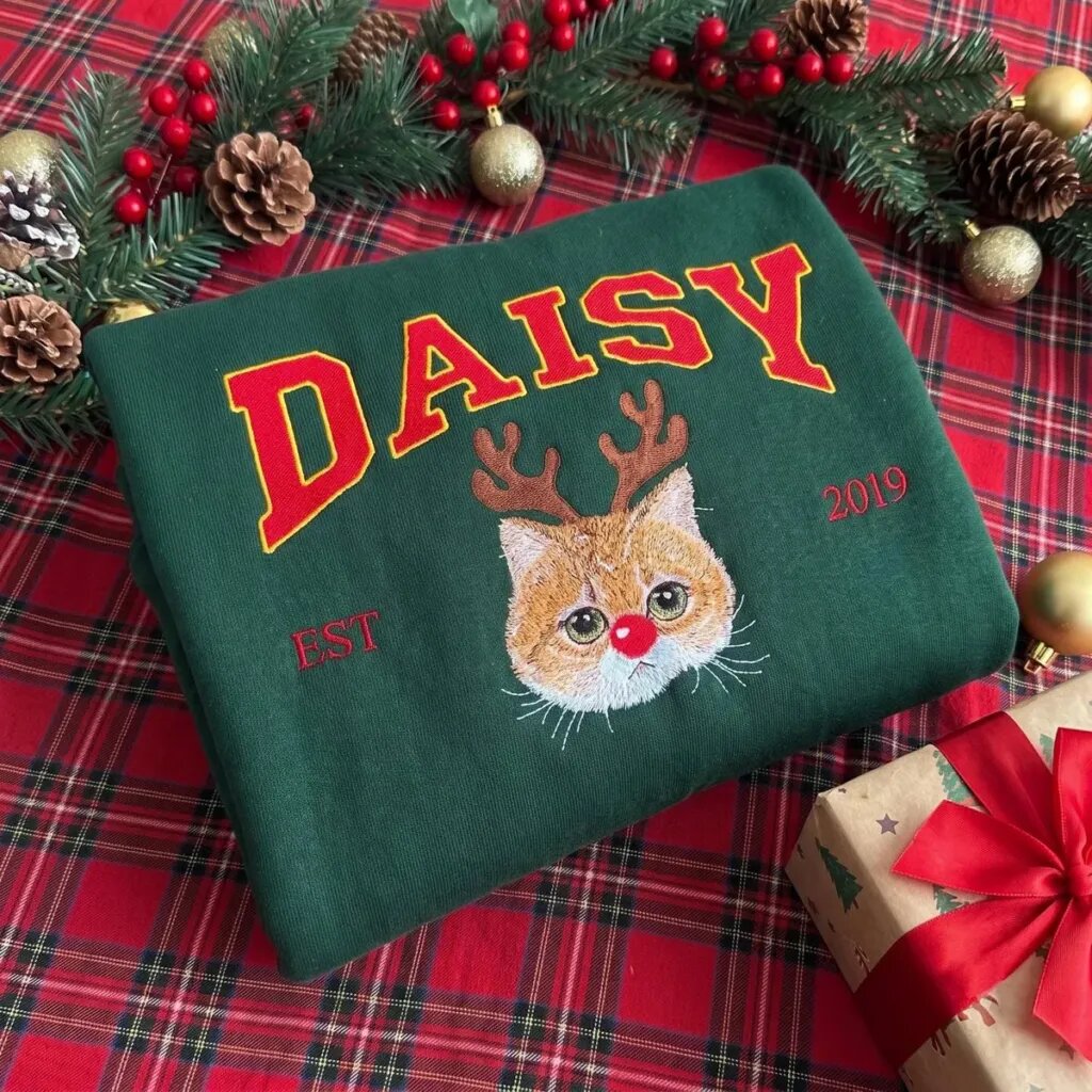 Personalized cat Christmas sweatshirt in Forest Green with DAISY in red varsity letters, an embroidered cat portrait with reindeer antlers and a red nose, and EST 2019