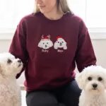 Maroon sweatshirt with embroidered portraits of two white dogs named Ruby and Max, worn next to the real dogs