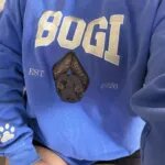 Woman wearing a blue custom dog crewneck with the name BOGI, an embroidered dog face, EST 2026 and a paw print on the sleeve