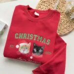Red crewneck with embroidered cat portraits, Santa hats and gold Merry Christmas script, styled with a straw mat