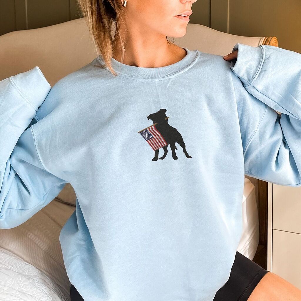 Woman in a Light Blue crewneck sweatshirt with a black pit bull holding a USA flag centered on the chest