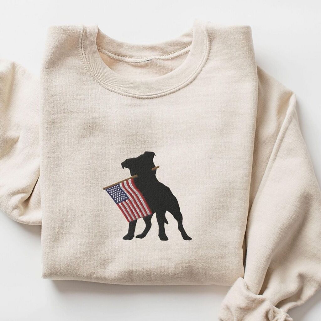 Sand crewneck sweatshirt laid flat with a rolled sleeve, embroidered with a pit bull silhouette holding a USA flag
