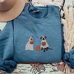 Indigo Blue crewneck sweatshirt laid flat on a patterned throw with a felt hat and pampas grass, embroidered with a pit bull and a black lab in ghost sheets holding jack-o'-lantern buckets