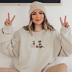 Smiling woman in a beanie making peace signs in a sand crewneck sweatshirt, embroidered with a pit bull and a black lab in ghost sheets holding jack-o'-lantern buckets