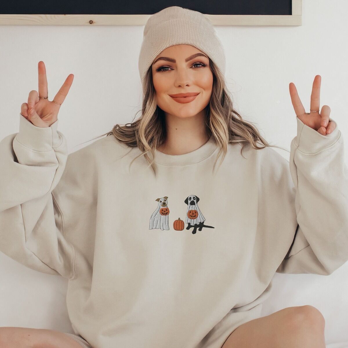 Pitbull and Black Lab Sweatshirt - worn in sand Smiling woman in a beanie making peace signs in a sand crewneck sweatshirt, embroidered with a pit bull and a black lab in ghost sheets holding jack-o'-lantern buckets