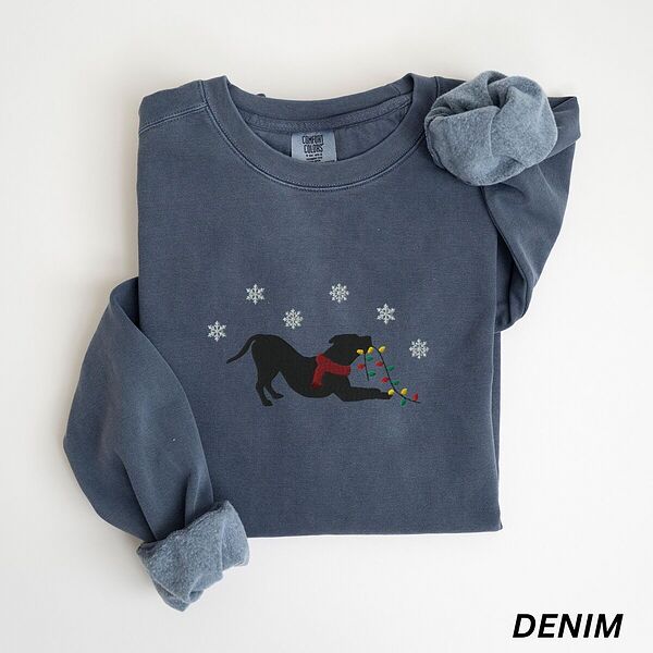 Denim crewneck sweatshirt laid flat with a rolled cuff, embroidered with a black pitbull silhouette in a play bow with a red scarf, holding a string of Christmas lights under white snowflakes