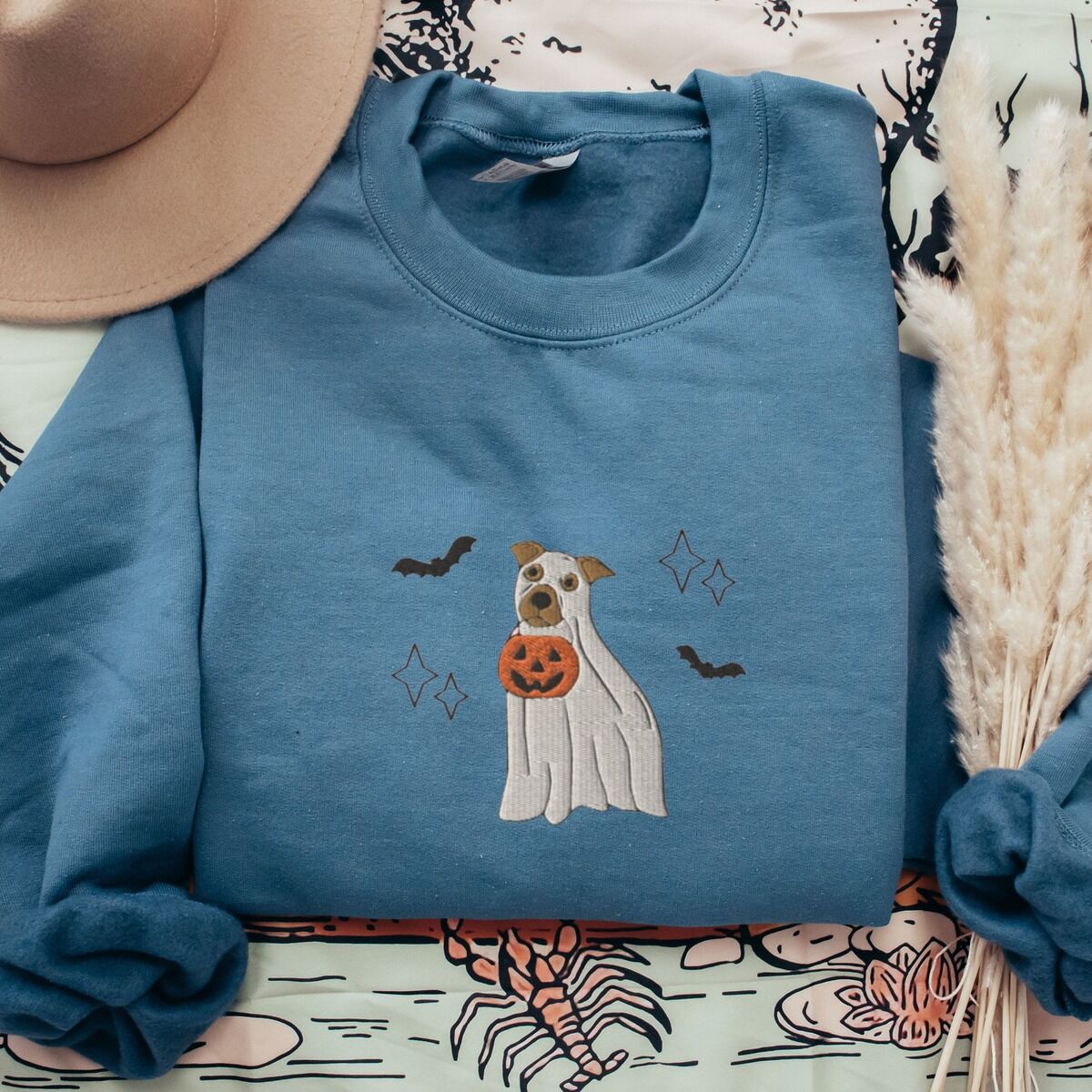 Pitbull Ghost Sweatshirt – Indigo Blue Indigo Blue crewneck sweatshirt laid flat on a patterned throw with a felt hat and pampas grass, embroidered with a pit bull in a ghost sheet holding a jack-o'-lantern bucket between bats and sparkles