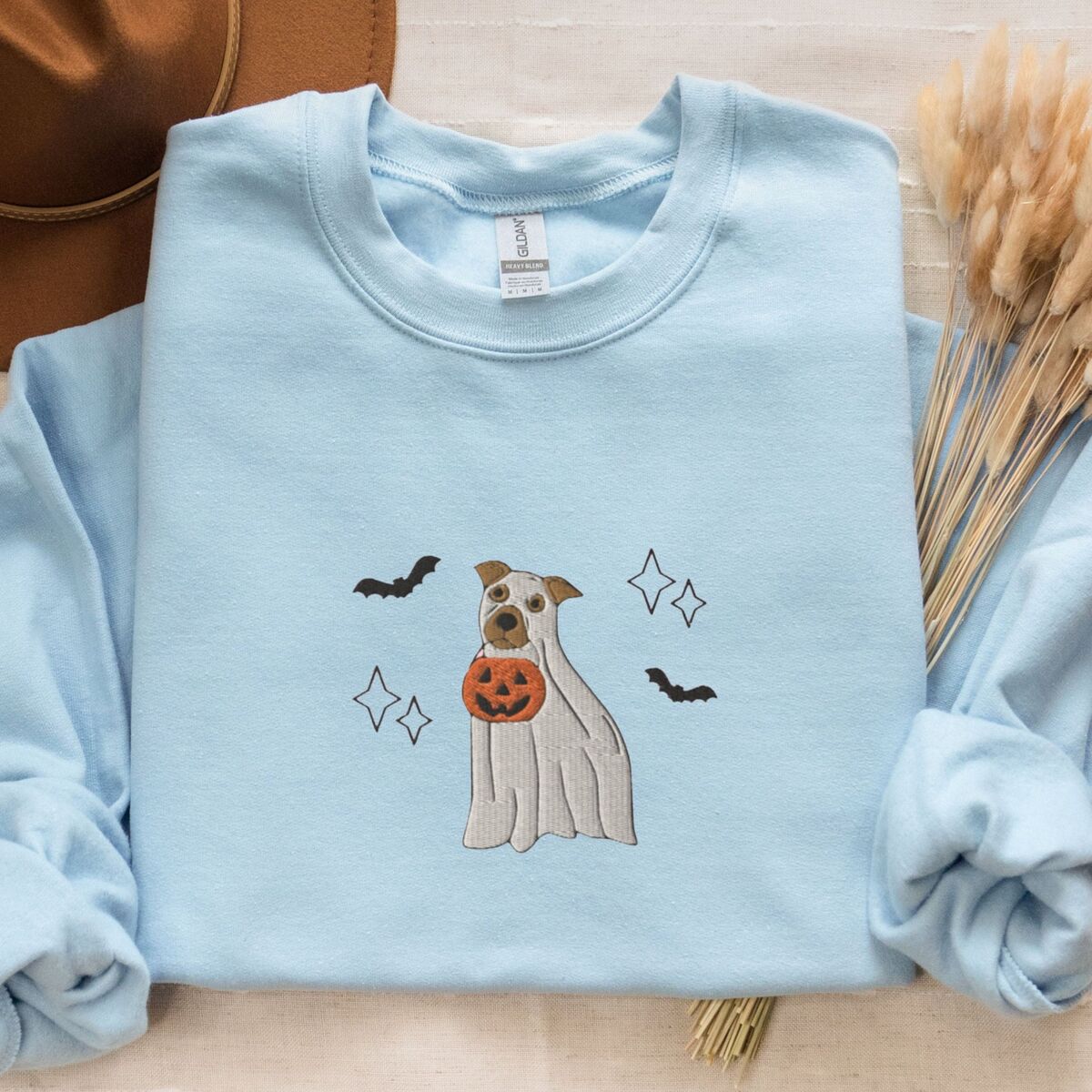 Pitbull Ghost Sweatshirt - Light Blue Light Blue crewneck sweatshirt laid flat with a brown hat and dried wheat, embroidered with a pit bull in a ghost sheet holding a jack-o'-lantern bucket between bats and sparkles