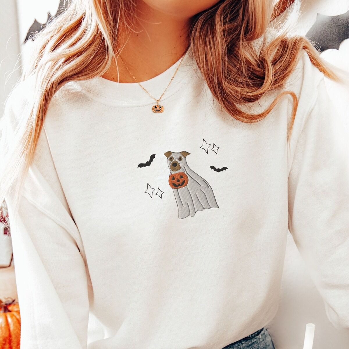 Pitbull Ghost Sweatshirt - worn Woman with long hair and a pumpkin necklace in a cream crewneck sweatshirt, embroidered with a pit bull in a ghost sheet holding a jack-o'-lantern bucket between bats and sparkles