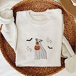 Sand crewneck sweatshirt folded in a woven basket, embroidered with a pit bull in a ghost sheet holding a jack-o'-lantern bucket between bats and sparkles