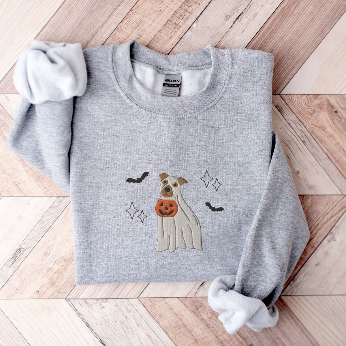Pitbull Ghost Sweatshirt - Sport Grey Sport Grey crewneck sweatshirt laid flat on a chevron wood floor, embroidered with a pit bull in a ghost sheet holding a jack-o'-lantern bucket between bats and sparkles
