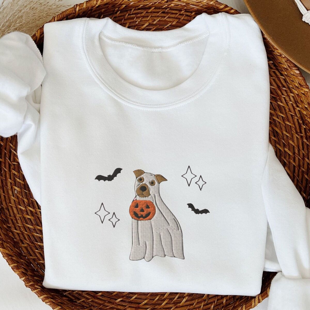 Pitbull Ghost Sweatshirt - White White crewneck sweatshirt folded in a woven basket next to a felt hat, embroidered with a pit bull in a ghost sheet holding a jack-o'-lantern bucket between bats and sparkles