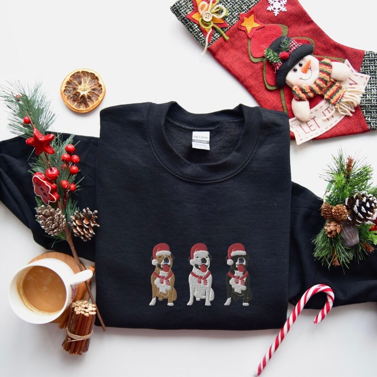 Pitbull Santa Hat Sweatshirt – Black Black crewneck sweatshirt laid flat with a Christmas stocking, candy cane and coffee, embroidered with three sitting pitbulls, fawn, white and black, in red Santa hats and red scarves