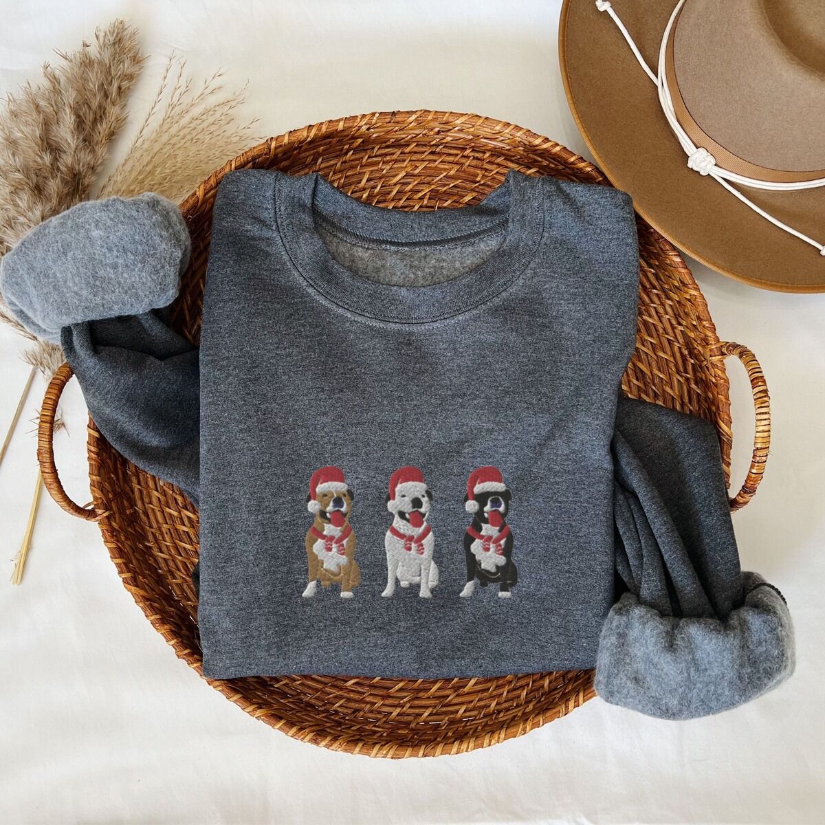 Pitbull Santa Hat Sweatshirt – Dark Heather Dark Heather crewneck sweatshirt folded in a woven basket, embroidered with three sitting pitbulls, fawn, white and black, in red Santa hats and red scarves