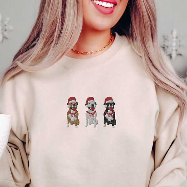 Smiling woman in a Sand crewneck sweatshirt embroidered with three sitting pitbulls, fawn, white and black, in red Santa hats and red scarves