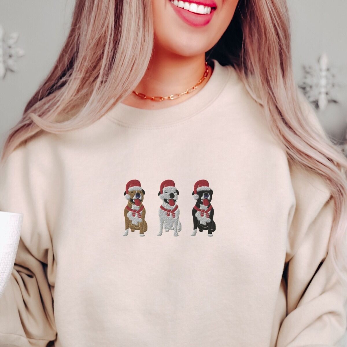 Pitbull Santa Hat Sweatshirt – Sand Smiling woman in a Sand crewneck sweatshirt embroidered with three sitting pitbulls, fawn, white and black, in red Santa hats and red scarves