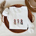 White crewneck sweatshirt folded in a woven basket, embroidered with three sitting pitbulls, fawn, white and black, in red Santa hats and red scarves