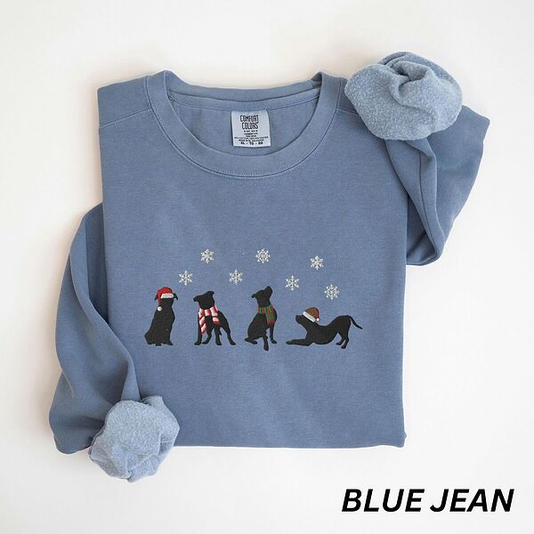 Blue Jean crewneck sweatshirt laid flat with a rolled cuff, embroidered with four black pitbull silhouettes in a Santa hat, striped scarves and a knit hat under snowflakes