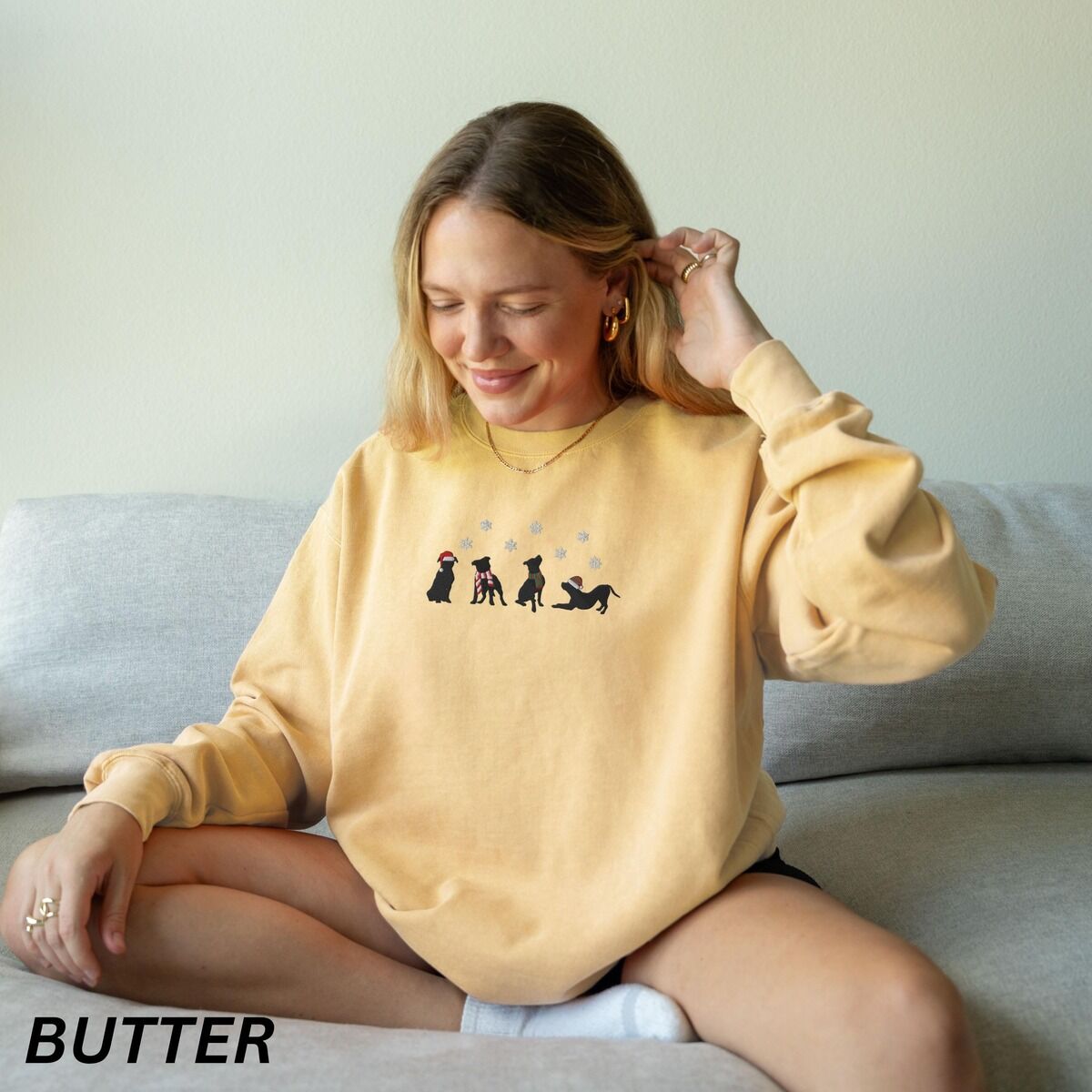 Pitbull Silhouette Christmas Sweatshirt – Butter Woman sitting on a sofa in a Butter crewneck sweatshirt embroidered with four black pitbull silhouettes in a Santa hat, striped scarves and a knit hat under snowflakes