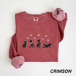 Crimson crewneck sweatshirt laid flat with a rolled cuff, embroidered with four black pitbull silhouettes in a Santa hat, striped scarves and a knit hat under snowflakes