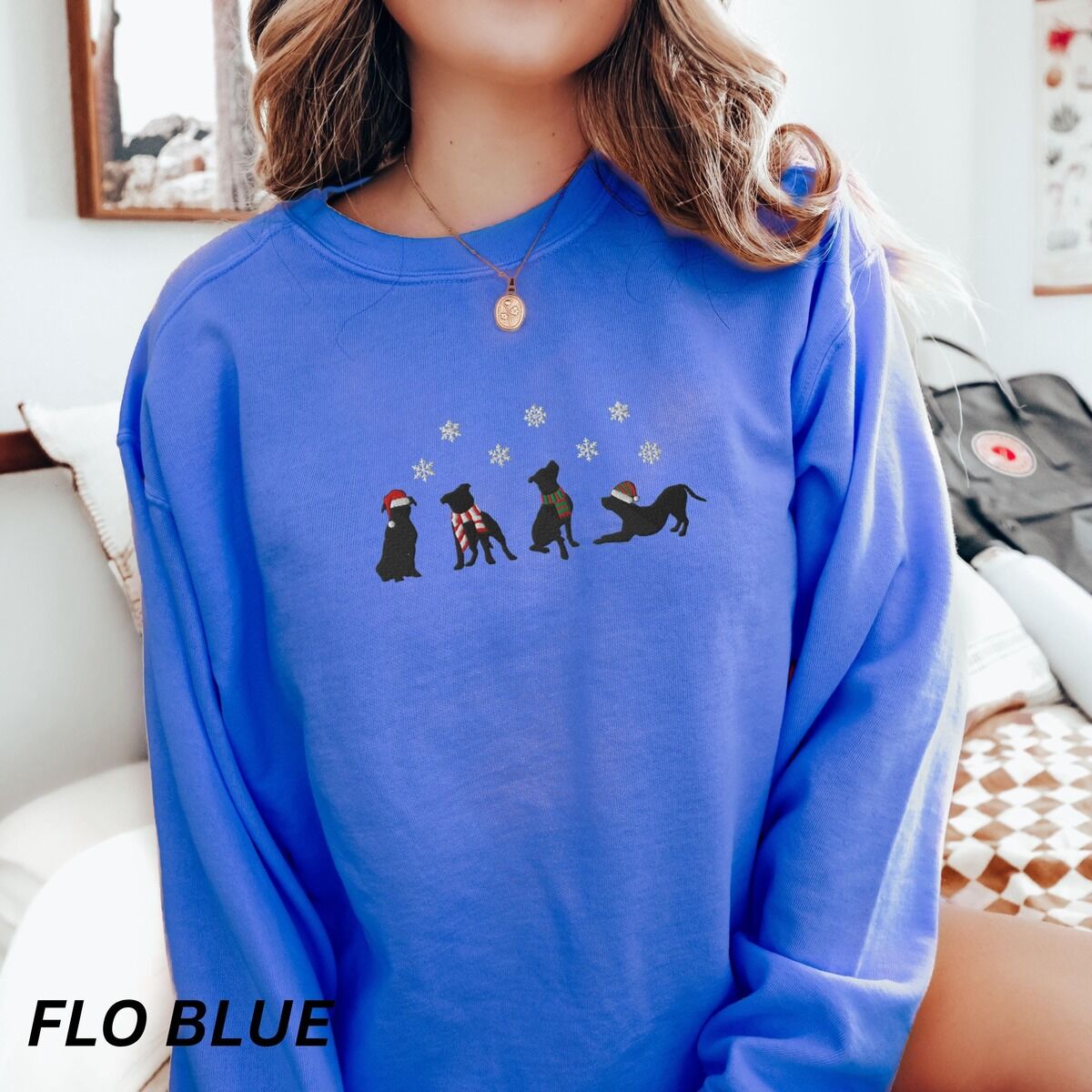 Pitbull Silhouette Christmas Sweatshirt – Flo Blue Woman in a Flo Blue crewneck sweatshirt and a pendant necklace, embroidered with four black pitbull silhouettes in a Santa hat, striped scarves and a knit hat under snowflakes