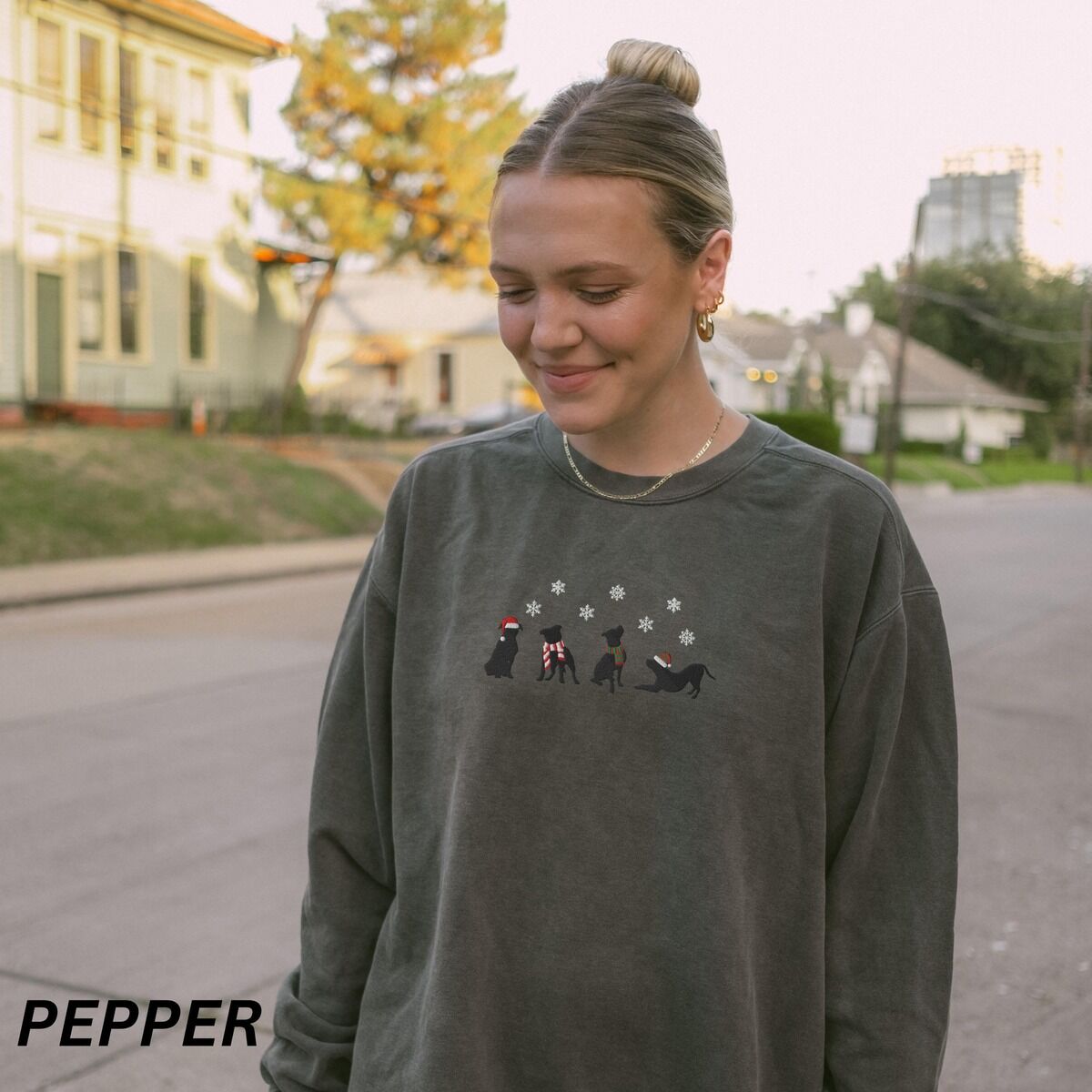Pitbull Silhouette Christmas Sweatshirt – Pepper Woman outdoors on a street in a Pepper crewneck sweatshirt embroidered with four black pitbull silhouettes in a Santa hat, striped scarves and a knit hat under snowflakes