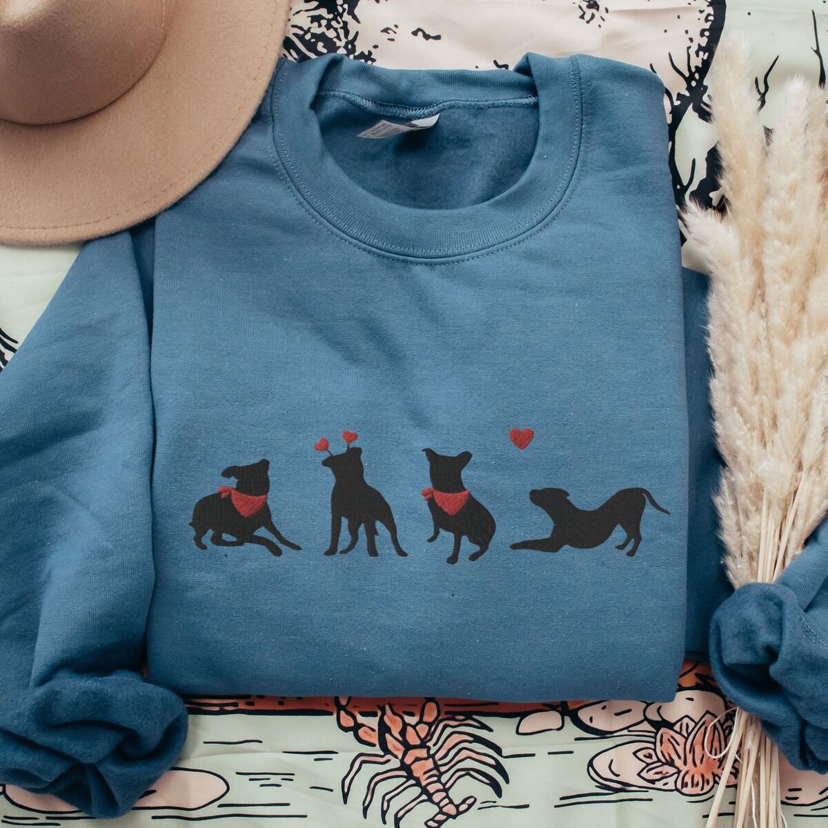 Pitbull Valentine Sweatshirt – Indigo Blue Indigo Blue crewneck sweatshirt laid flat with a tan hat and dried pampas, embroidered with four black pitbull silhouettes with red bandanas, a red heart headband and a floating red heart