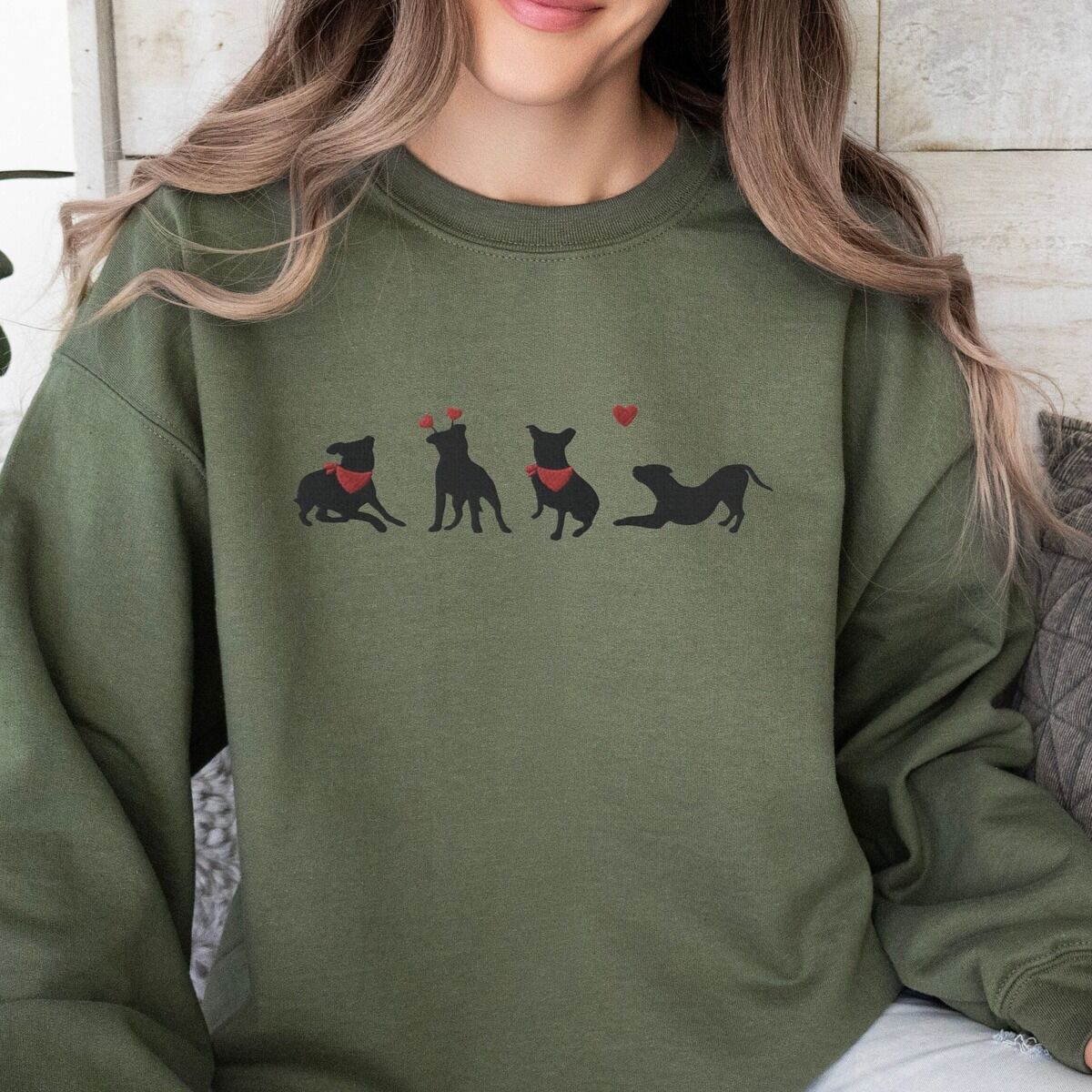 Pitbull Valentine Sweatshirt – Military Green Smiling woman in a Military Green crewneck sweatshirt embroidered with four black pitbull silhouettes with red bandanas, a red heart headband and a floating red heart