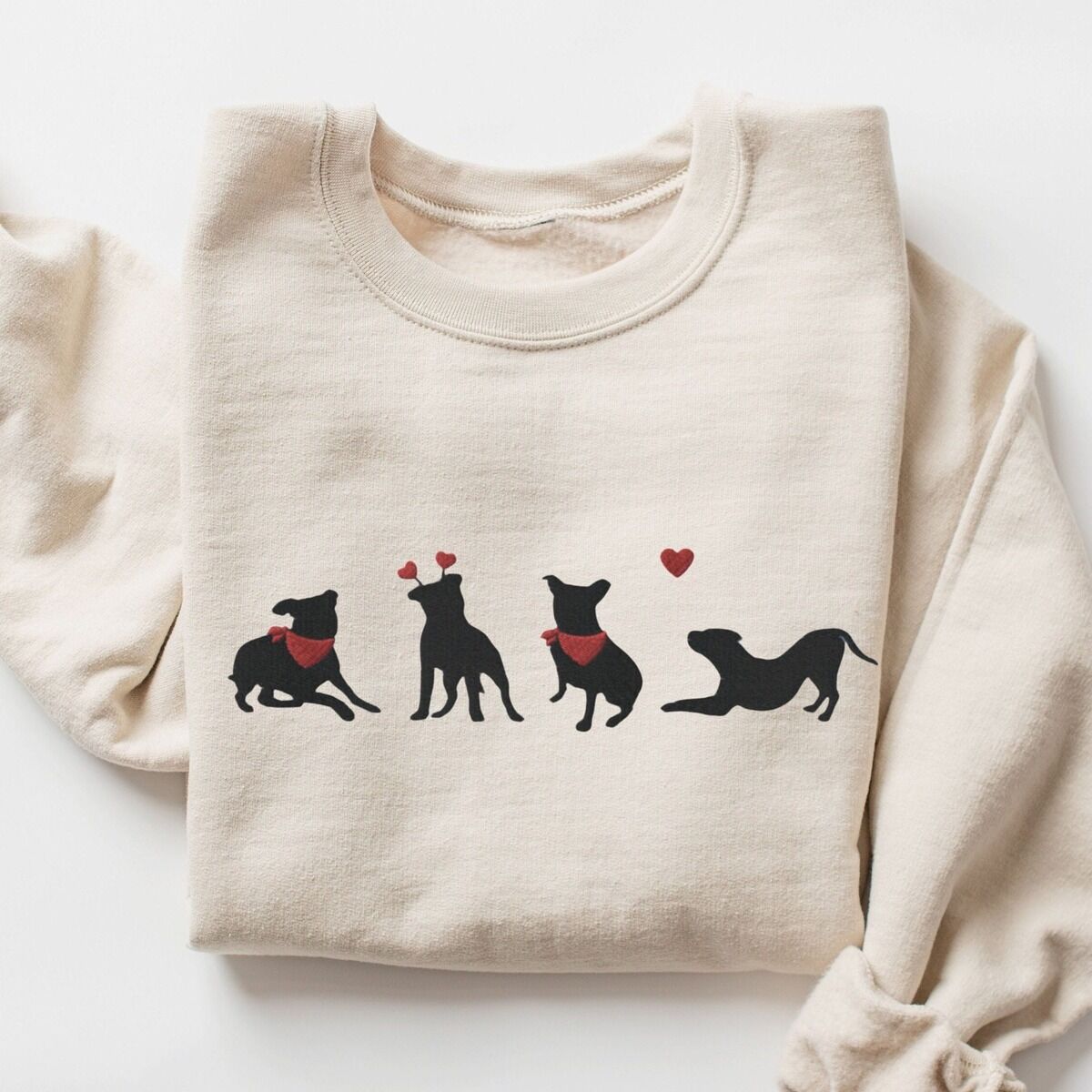 Pitbull Valentine Sweatshirt – Sand Sand crewneck sweatshirt laid flat, embroidered with four black pitbull silhouettes with red bandanas, a red heart headband and a floating red heart