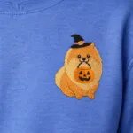Close-up of the embroidered Pomeranian in a witch hat holding a jack-o'-lantern bucket on the left chest of a Heather Deep Royal sweatshirt