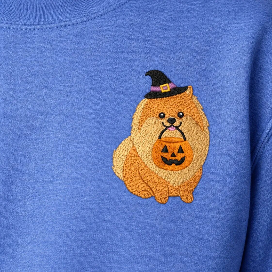 Pomeranian Halloween Embroidery – Left Chest Close-up Close-up of the embroidered Pomeranian in a witch hat holding a jack-o'-lantern bucket on the left chest of a Heather Deep Royal sweatshirt