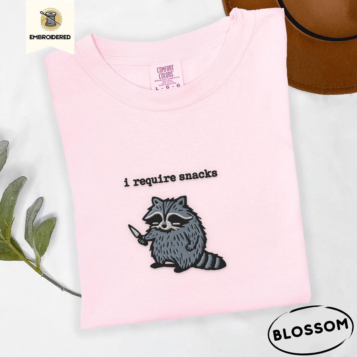 Raccoon Shirt – Blossom Blossom Comfort Colors T-shirt embroidered with a feral raccoon holding a little knife under the words i require snacks embroidered on the chest