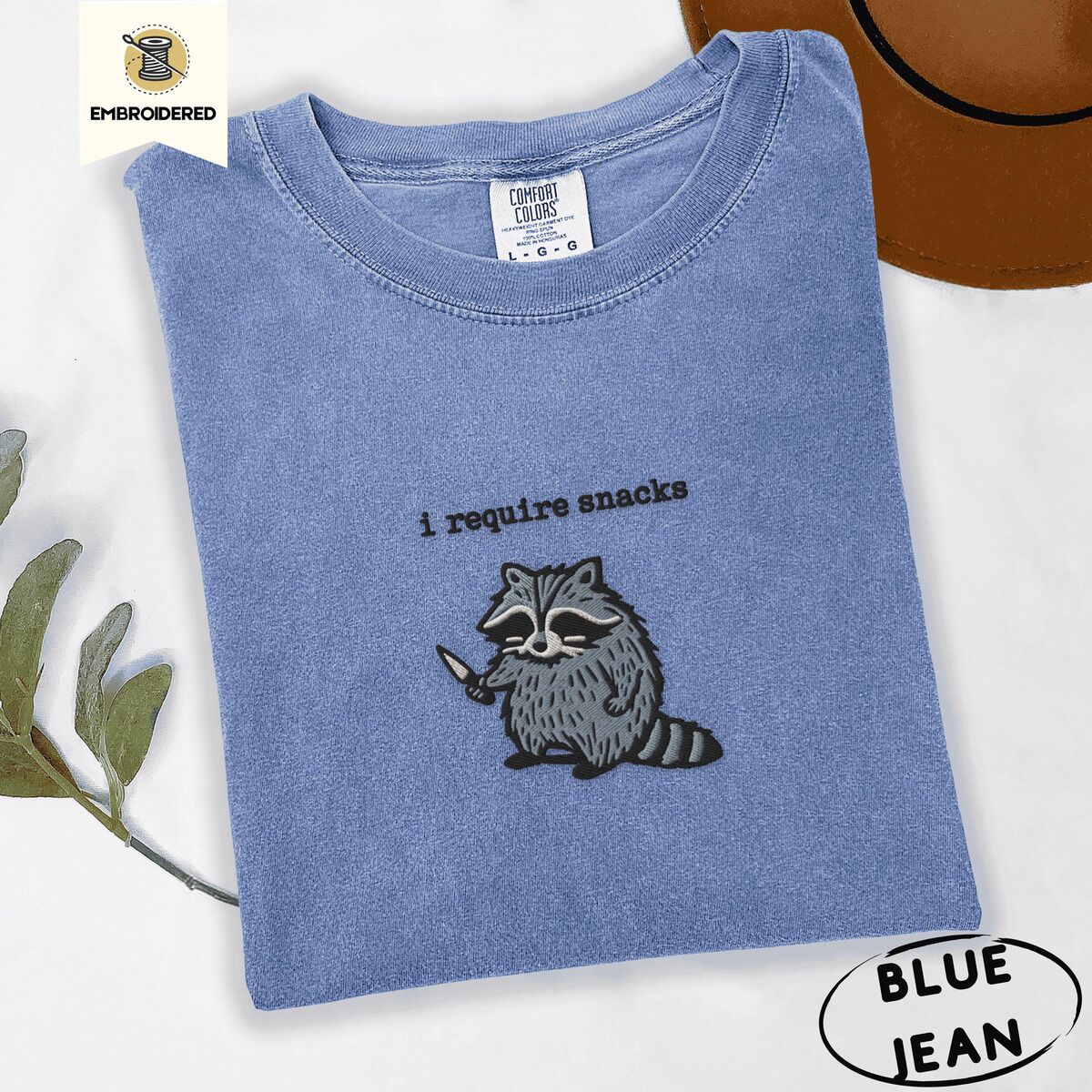 Raccoon Shirt – Blue Jean Blue Jean Comfort Colors T-shirt embroidered with a feral raccoon holding a little knife under the words i require snacks embroidered on the chest