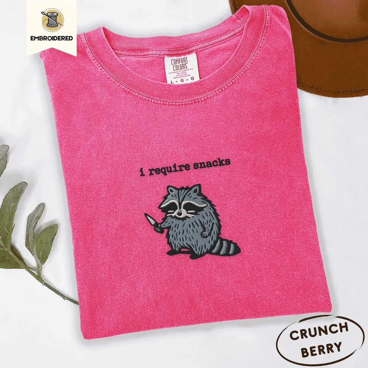 Raccoon Shirt – Crunchberry Crunchberry Comfort Colors T-shirt embroidered with a feral raccoon holding a little knife under the words i require snacks embroidered on the chest